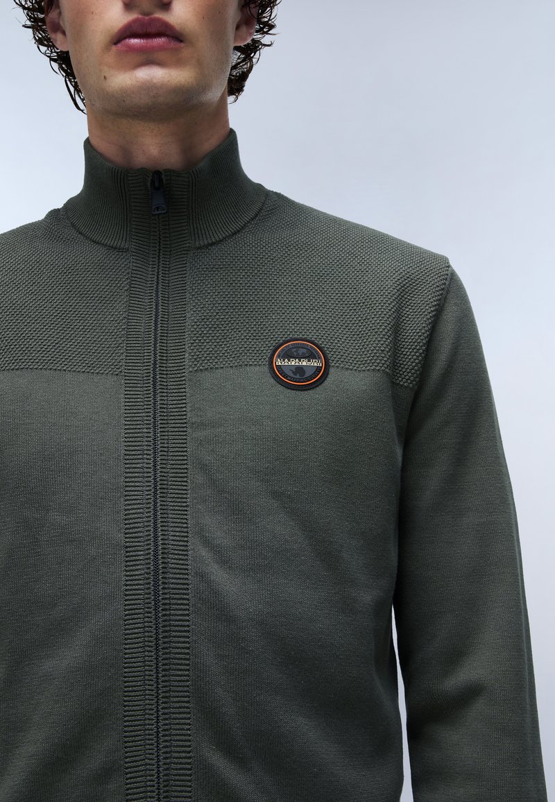 Gilet zippé kaki avec un col montant, panneau supérieur texturé et un écusson circulaire. Accents côtelés le long de la fermeture éclair. Matière en tricot doux.