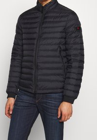 Man med en svart vadderad puffjacka med dragkedja fram och dragkedjefickor på sidan, i kombination med mörkblå jeans.
