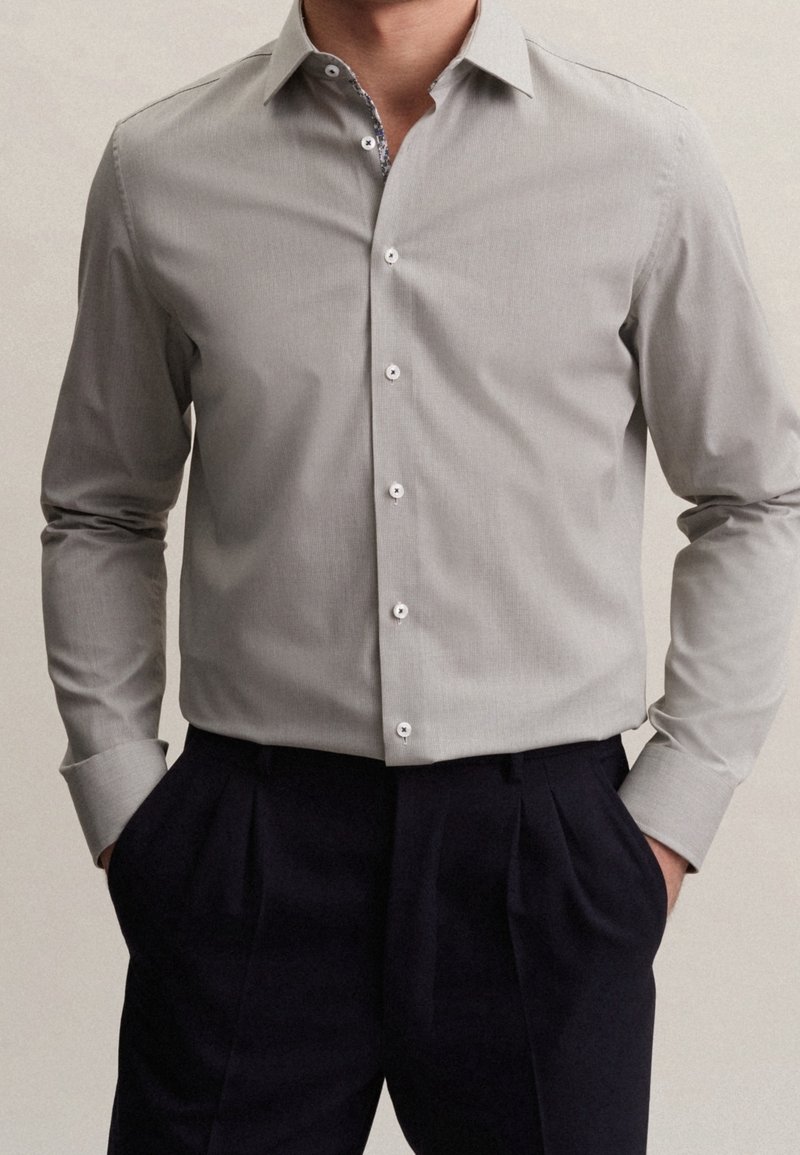 Grå knapp-up skjorta med en subtil textur, spetsig krage och vita knappar. Bärs med mörka veckade byxor.