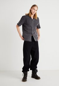 Cămașă din denim negru decolorat, cu mâneci scurte și închidere cu nasturi, asortată cu pantaloni negri largi și cizme maro cu șireturi.
