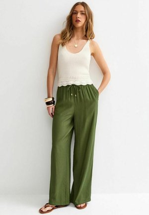 Vrouw draagt een mouwloze crèmekleurige gebreide top, olijfgroene wijde broek, bruine sandalen en zwarte en beige armbanden, staand tegen een witte muur.