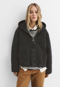 Geacă neagră cu glugă, cu fermoar pe față și două buzunare, stratificată peste un tricou cu dungi și o cămașă albă cu nasturi, asortată cu pantaloni maro.