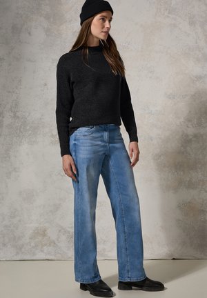 Pull en maille noir texturé, jean large bleu clair, et bottines noires. Le modèle porte un bonnet noir assorti, se tenant devant un fond neutre.