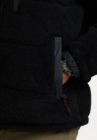 Person som bär en svart fleecejacka med handen i fickan, som visar en klocka med grön urtavla och svart rem.