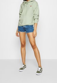 Svetle zelená mikina s kapucí a šňůrkou, přední kapsa; modré džíny se třepeným lemem; olivově zelené tenisky s bílými akcenty a tkaničkami.