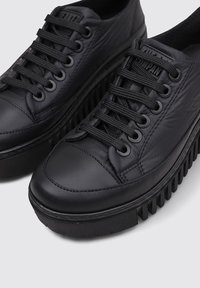 Zapatillas negras con un empeine texturizado, punta redonda y suela gruesa con estrías. Presentan cordones y ojales negros, con un sutil logo en la lengua.