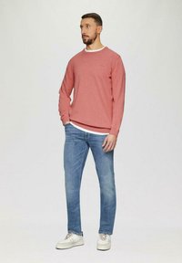 Rotes Sweatshirt mit Rundhalsausschnitt, über einem weißen T-Shirt getragen, kombiniert mit blauen Jeans und weißen Sneakers, vor einem hellen Hintergrund stehend.