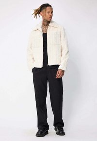 Un homme avec des dreadlocks portant une veste blanche en peluche, une chemise noire, un pantalon noir et des chaussures noires, se tenant devant un fond blanc uni.