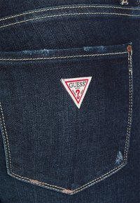 Tummansinisiä farkkuja, joissa on takataskussa kolmion muotoinen punavalkoinen 'GUESS' -logo, näkyvää tikkausta ja hieman kuluneita yksityiskohtia.