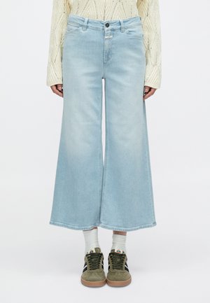 Hellblaue Jeans mit weitem Bein aus Denim, mit mittlerer Waschung, hoher Taille und einem Knopfverschluss. Kombiniert mit grünen Sneakers und weißen Socken.