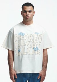 Fehér, oversize póló, amelyen a "BROKEN HEART CLUB" felirat olvasható vastag betűkkel, világoskék szívgrafikákkal kiemelve. 100% pamut anyag, laza szabású.