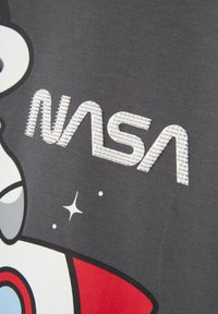 A szürke szöveten fehér NASA logó látható, texturált felülettel, stilizált űrtematikájú grafikákkal és piros kiegészítőkkel.