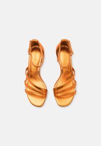 Fényes narancssárga, pántos magassarkú szandál, bokapánttal és négyzet alakú orral, felülről nézve, fehér háttéren.