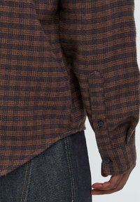 Camisa de tela a cuadros en marrón y azul marino con una textura suave, con puños abotonados y un dobladillo ligeramente curvado. Combinada con vaqueros oscuros.