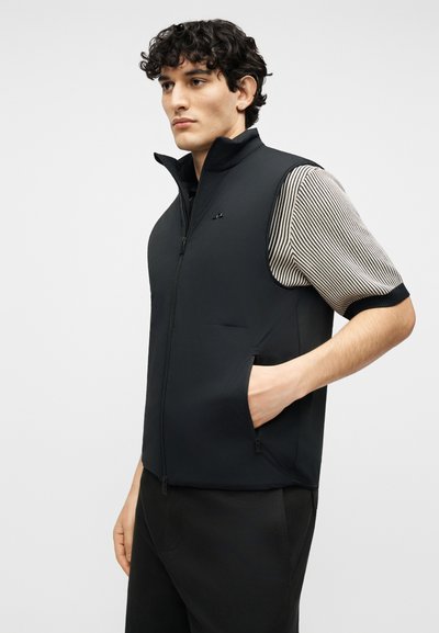 Νέος με σγουρά μαλλιά που φοράει μαύρο γιλέκο με φερμουάρ πάνω από κοντομάνικο ριγέ πουκάμισο, με το χέρι στην τσέπη του γιλέκου, κοιτάζει μπροστά.