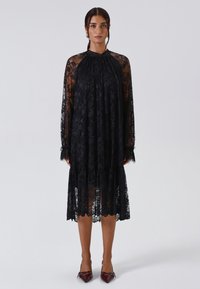 Vestito nero in pizzo con maniche lunghe, colletto alto e trama trasparente. Presenta intricate fantasie floreali e un orlo a strati, abbinato a tacchi bordeaux.