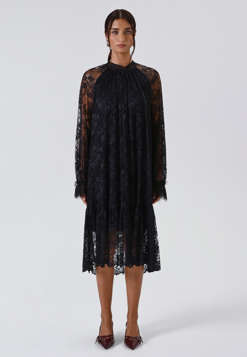 Vestito nero in pizzo con maniche lunghe, colletto alto e trama trasparente. Presenta intricate fantasie floreali e un orlo a strati, abbinato a tacchi bordeaux.