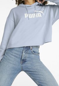 Persoon in een lichtblauwe cropped Puma-hoodie en hoge blauwe denim jeans, poseert met één arm omhoog bij het hoofd.