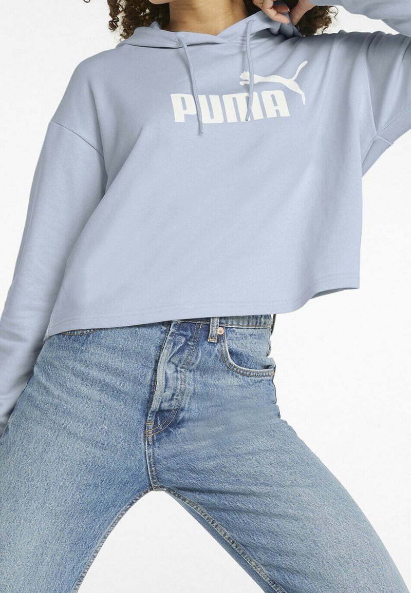 Persoon in een lichtblauwe cropped Puma-hoodie en hoge blauwe denim jeans, poseert met één arm omhoog bij het hoofd.