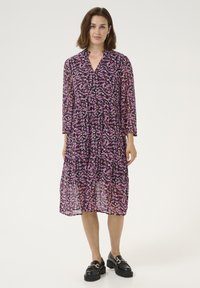 Purple semi-sheer midi dress with a floral pattern, a v-neckline, and three-quarter sleeves. Finished with a tiered hem and worn with black loafers.