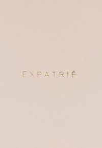 Miękka beżowa skóra o gładkiej fakturze, z wytłoczonym złotym napisem "EXPATRIÉ" umieszczonym na środku powierzchni.