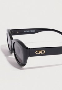 Μαύρα υπερμεγέθη γυαλιά ηλίου με γυαλιστερό φινίρισμα. Διαθέτουν στρογγυλούς φακούς και χρυσές λεπτομέρειες υλικού στο πλάι. Το κείμενο γράφει "ANNA FIELD."