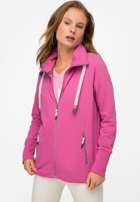 Felpa rosa con zip e colletto alto, cordini grigi e due tasche anteriori con zip. Il tessuto sembra liscio ed elastico.