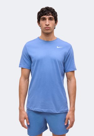Man staand met het gezicht naar voren, draagt een blauw Nike T-shirt en blauwe shorts, neutrale uitdrukking, effen lichte achtergrond.