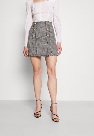 Woman wears a black and white houndstooth mini skirt with gold buttons, paired with a white long-sleeve top and black high-heeled sandals.