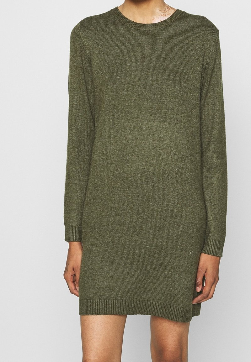 Personne portant une robe pull en maille vert olive à manches longues avec des poignets et un ourlet côtelés, debout devant un fond clair uni.