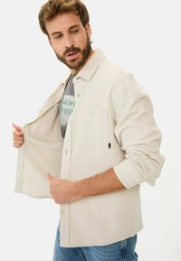 Lys beige knap-op skjorte med en frontlomme, tekstureret stof, åben krave og lange ærmer, lagd over en grafisk t-shirt.