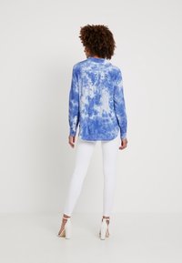 Chemise manches longues à tie-dye bleu avec un ourlet arrondi, des poignets boutonnés, et un jean skinny blanc. Le fond est uni et clair.