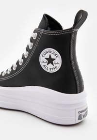 Svart høyhælte sneakers med en teksturert læroverdel, hvite sømmer og en fremtredende sirkulær logo på siden. Tykk hvit plattformsåle.
