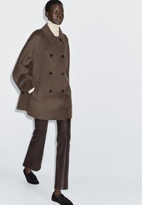 Brun dubbelknäppt ullrock med svarta knappar, kombinerad med bruna läderbyxor och svarta slip-on skor, uppvisad mot en enkel bakgrund.