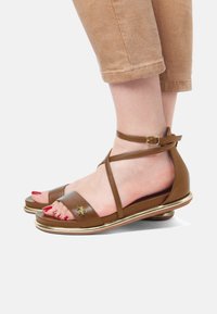 Ruske nahkasi sandalsit, joissa on nilkkahihna, kultaiset korostukset ja avonainen varvasmuoto. Hiukan kohotettu pohja, jossa on yksityiskohtaista ompelua päällisessä.