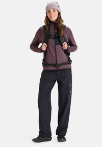 Paarse fleecejack met rits, zwarte broek met zakken, en rugzak, gedragen met een lichtpaarse gebreide muts en zwarte schoenen.