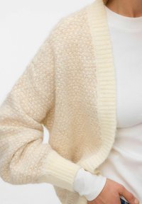 Krēmkrāsas teksturēts kardigans ar ribotu apkakli, mīkstu audumu, atvērtu priekšpusi un plašām piedurknēm, pārģērbts pār baltu pieguļošu topu.