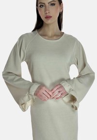 Top beige de manga larga con textura acanalada, puños anchos con volantes y escote redondeado. Textura suave y ajuste relajado. Manos colocadas en la cintura.