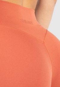 Růžovo-oranžový sportovní top s hladkým povrchem, který má vysoký límec a jemný detail loga na zadní straně. Je viditelné šití švů.