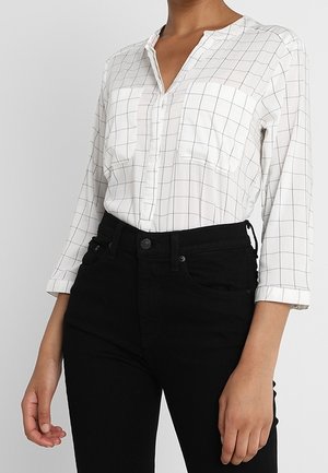 Kobieta w białej bluzce z długim rękawem z cienkim czarnym wzorem w kratkę, włożona do wysokich czarnych spodni, stojąca na jednolitym tle.