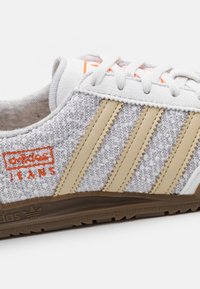 Šedé textilní tenisky s béžovými pruhy, bílými šňůrkami a oranžovým brandingem. Hnědá gumová podrážka má texturované detaily.