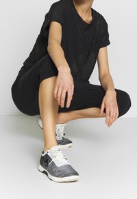 Schwarzes Mesh-T-Shirt mit kurzen Ärmeln, kombiniert mit schwarzen, verkürzten Jogginghosen. Graue und weiße Sportschuhe mit weißen Schnürsenkeln.