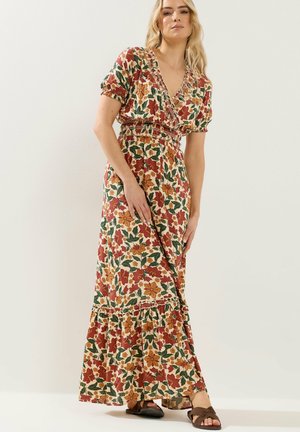 Blonde Frau in einem geblümten Maxikleid mit roten, orangefarbenen und grünen Blumen, posiert mit einer Hand, die den Rock hält, braune Sandalen sichtbar.