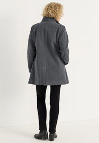 Grau melierter Wollmischungenmantel mit hohem Kragen, übergroßen Ärmeln und geradem Schnitt. Getragen über schwarzen Slim-Fit-Hosen und Ankle Boots.