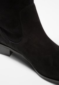 Bottine à cheville en daim noir avec une silhouette droite, un bout rond et un petit talon bloc. Texture lisse avec des détails de couture décorative.