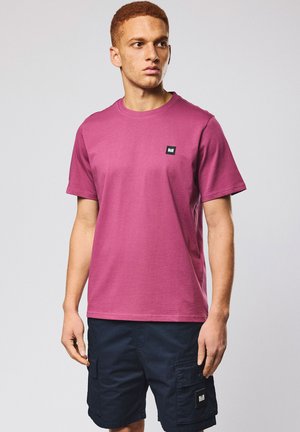 Mann mit kurzen lockigen Haaren, der ein dunkelrosa T-Shirt und marineblaue Cargo-Shorts trägt, vor einem einfarbigen hellen Hintergrund stehend.