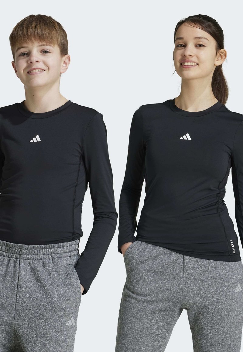 Zwarte lange mouwen sportshirts met wit logo, gecombineerd met grijze gestructureerde joggers. Beide kledingstukken hebben een aansluitende pasvorm.