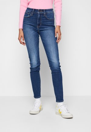 Vaqueros ajustados de tiro alto en azul marino oscuro con un leve desvanecimiento, diseño de cinco bolsillos y costuras en contraste, combinados con zapatillas blancas.