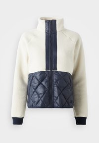 Flis jakna s bijelim gornjim dijelom i mornarsko plavim k quilted donjim dijelom. Ima visoki ovratnik, prednji zatvarač i rebraste manžete.