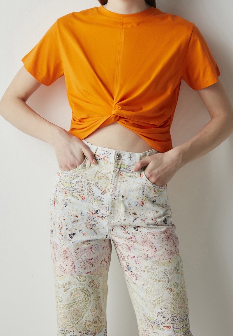 Personne portant un crop top noué orange et un pantalon taille haute clair à motif paisley multicolore, mains dans les poches.